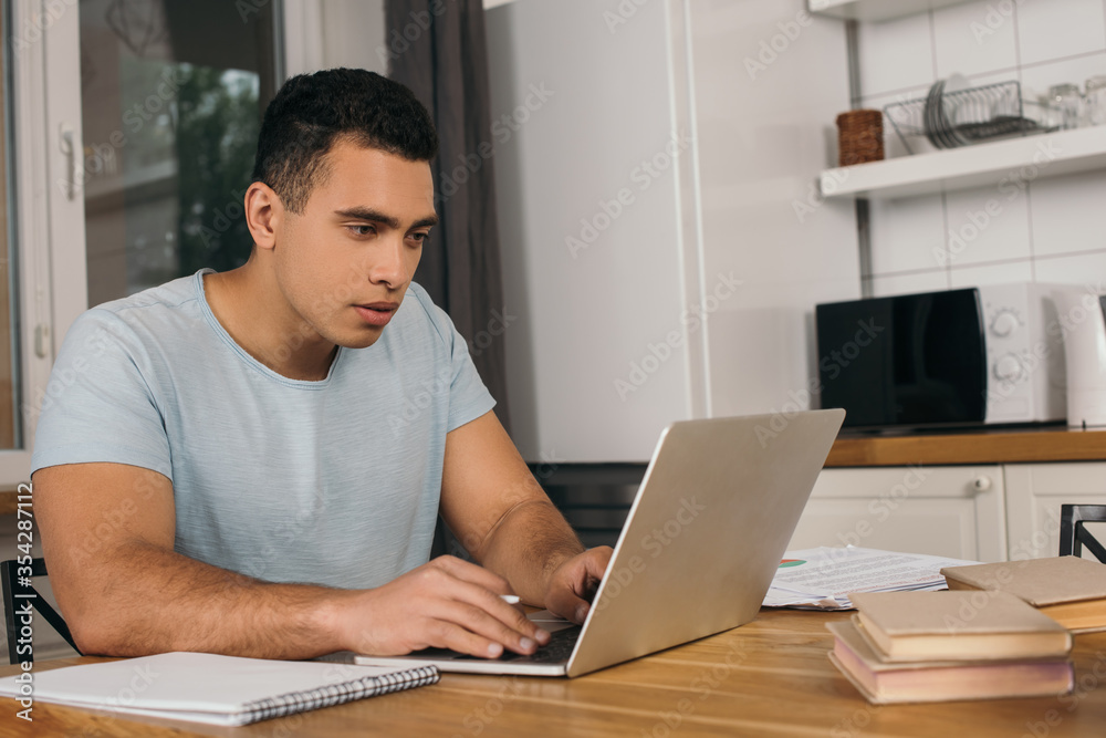 handsome mixed race freelancer using laptop near books and notebook handsome mixed race freelancer using laptop near books and notebook