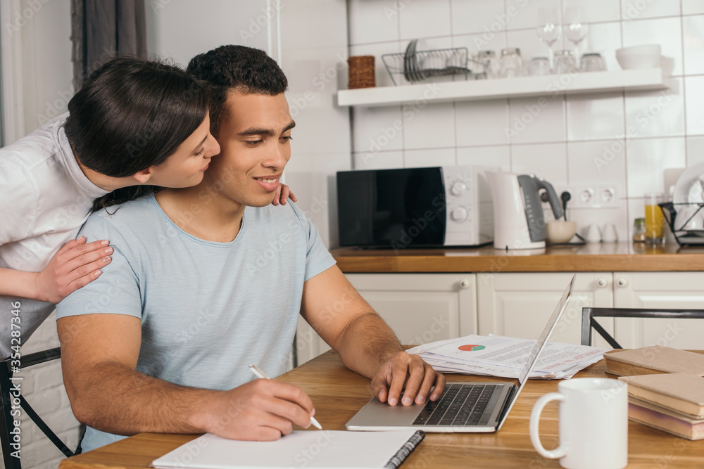 cheerful woman kissing cheek of happy mixed race boyfriend holding pen near notebook and laptop, online study concept cheerful woman kissing cheek of happy mixed race boyfriend holding pen near notebook and laptop, online study concept
