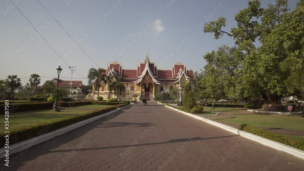 Tourists on footpath against famous hall by trees during sunny day - Vientiane, Laos