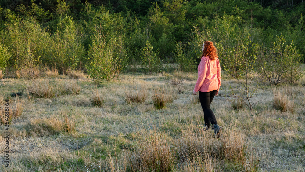 Young woman walking through a meadow carrying a camera, in late evening light.