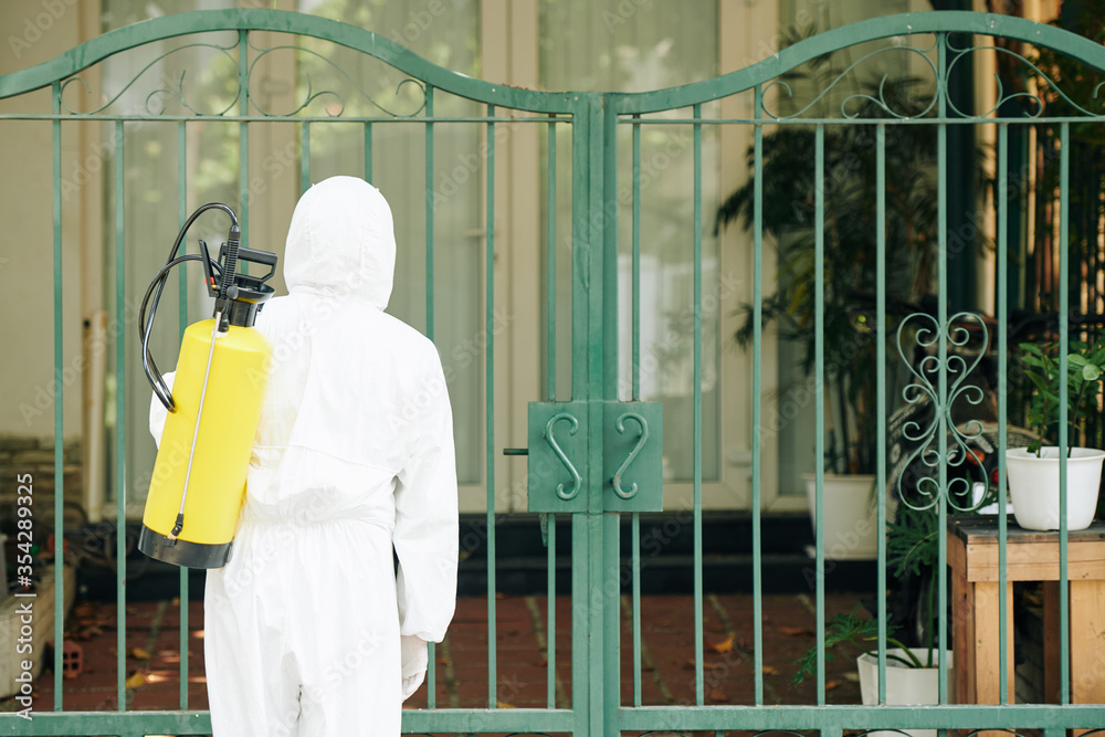 Disinfection service worker in protective suit spraying house gates, view from the back Disinfection service worker in protective suit spraying house gates, view from the back