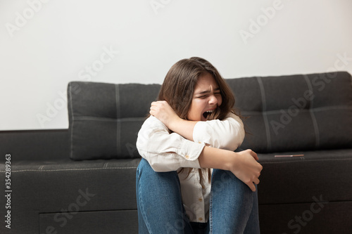 young brunette woman looks desperate crying loud while sitting near sofa in her appartment, emotional nervous breakdown concept photo