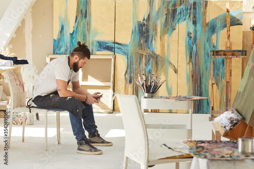 Full length side view at contemporary bearded artist using smartphone while sitting on chair in art studio with abstract paintings in background, copy space photo