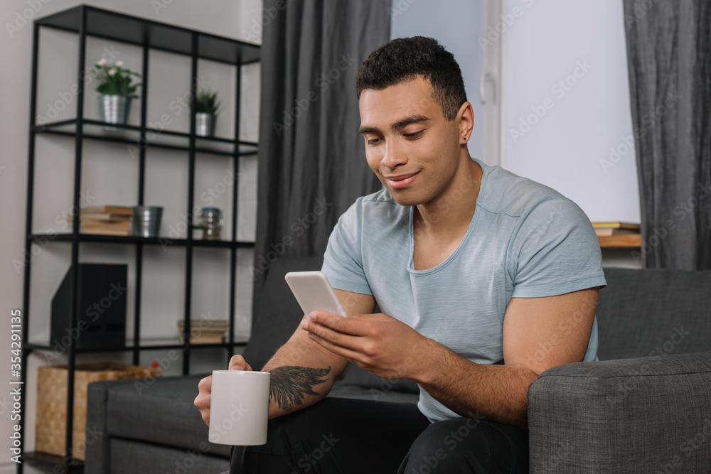 happy mixed race man with tattoo holding cup and using smartphone happy mixed race man with tattoo holding cup and using smartphone