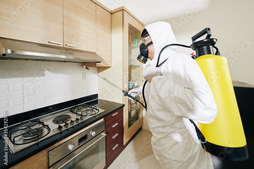 Infection prevention and control of epidemic concept: worker in white hazmat suit spaying detergent on kitchen cabinets