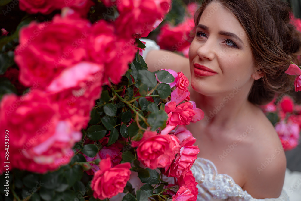 Naklejka premium Portrait of pretty bride dressed wedding white dress, make up and hairstyle posing at the background flowers roses wall at the morning with wind in hair.