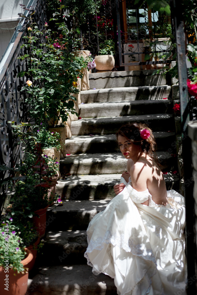 Alone luxury bride wearing stylish wedding white dress waiting in the stairs of the house with flowers
