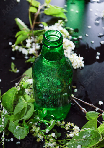Green glass bottles with flowers bird cherry and green leaves on light dark background with water drops