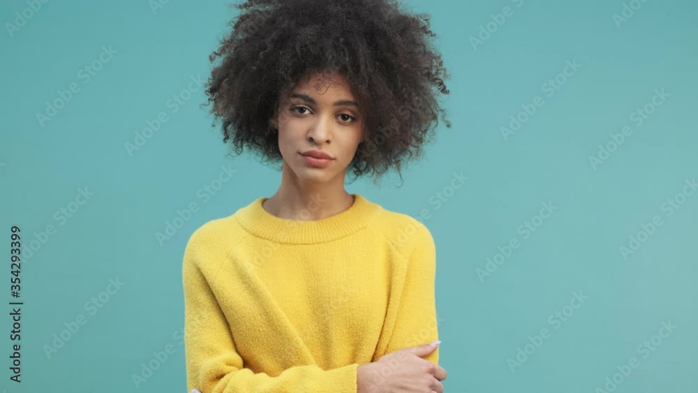A calm young african american woman with curly hair is looking to the camera standing with crossed hands isolated over blue wall background in studio