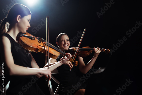 laughing female musicians playing on violins on dark stage with back light