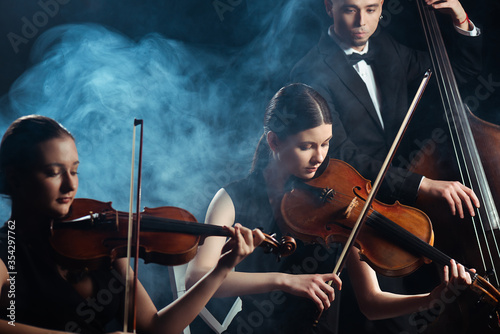 Tablou pe pânză trio of musicians playing on violins and contrabass on dark stage with smoke