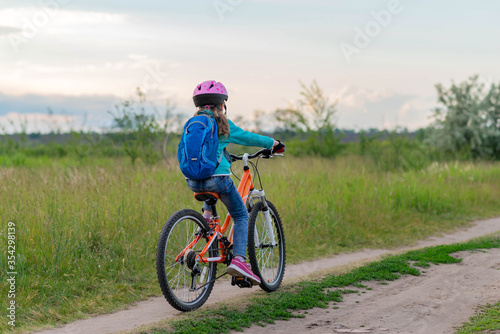 A little girl rides a bicycle on a country road along the meadow.