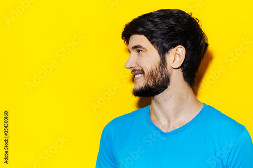 Studio profile portrait of young smiling guy in cyan shirt looking on empty space of yellow background.