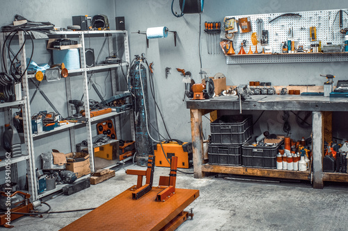 Garage, service area for disassembling, repairing motorcycles, car service station. Inside the workshop with large workbench, shelving, moto lift, tools kit for processing wrenches on the wall
