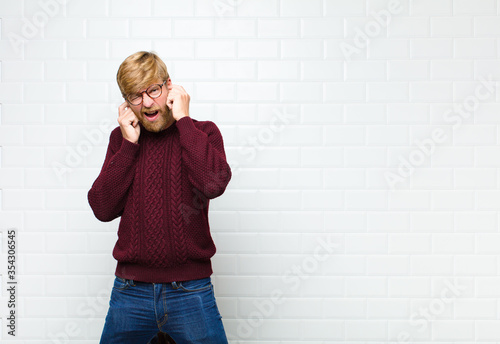 young blonde man looking angry, stressed and annoyed, covering both ears to a deafening noise, sound or loud music agaist vintage tiles wall photo