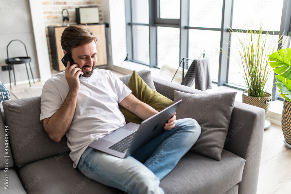 Young man using a laptop while talking on the phone sitting on the couch at home Young man using a laptop while talking on the phone sitting on the couch at home
