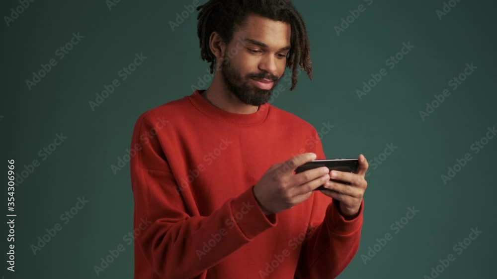A handsome young african american man is holding his smartphone horizontally standing isolated over green wall background in studio A handsome young african american man is holding his smartphone horizontally standing isolated over green wall background in studio