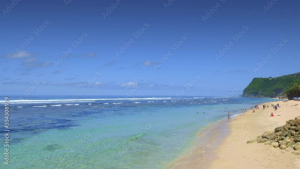 Turquoise colored tropical beach with small waves in transparent and clean water with people are walking and resting on bright yellow sand. High tide waves are crushing reef in background.