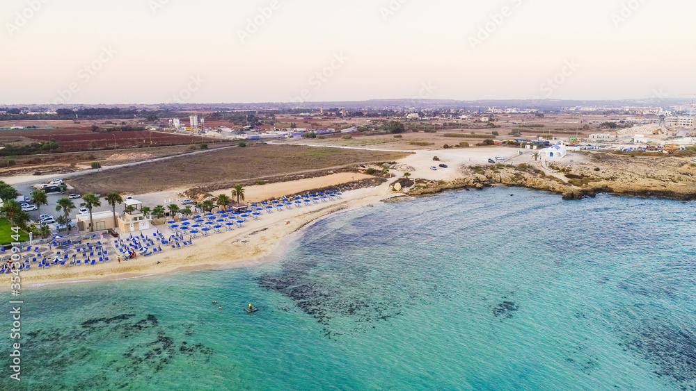 Aerial view of coastline sunset and landmark beach of Agia Thekla, Ayia Napa, Famagusta, Cyprus from above. Bird's eye skyline view of tourist attraction golden sand bay, islet, sunbeds in Ammochostos