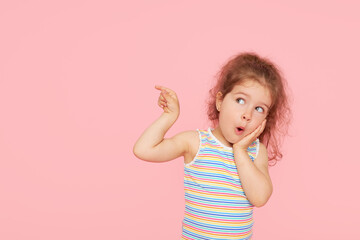 Portrait of surprised cute little toddler girl child over pink background. Looking at camera. Points hands to the left side. Advertising childrens products