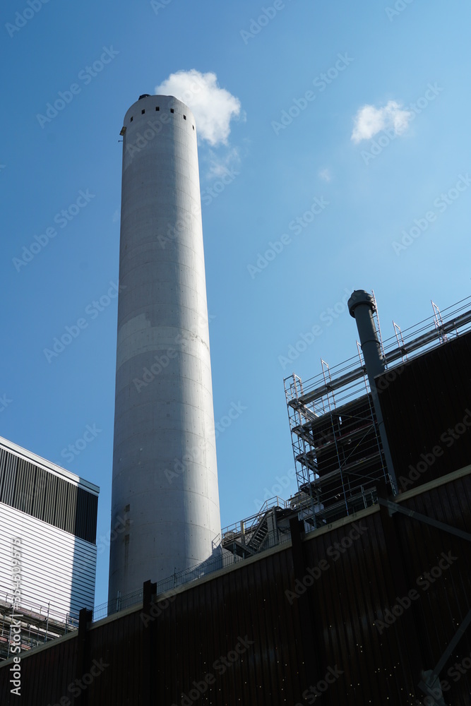 a tall chimney on an industrial building or factory surrounded by other structures indicating industrial environment. the chimney is emitting smoke. a tall chimney on an industrial building or factory surrounded by other structures indicating industrial environment. the chimney is emitting smoke.