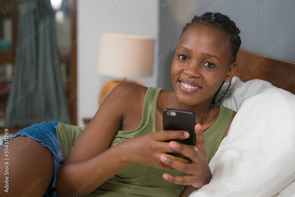 lifestyle home portrait of young happy and attractive black African American woman lying on bed using social media app in mobile phone relaxed and cheerful online dating