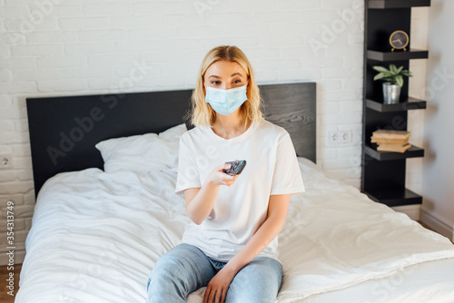 Young woman in medical mask holding remote controller while sitting on bed