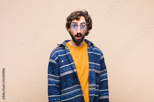 young crazy cool man feeling terrified and shocked, with mouth wide open in surprise against flat wall