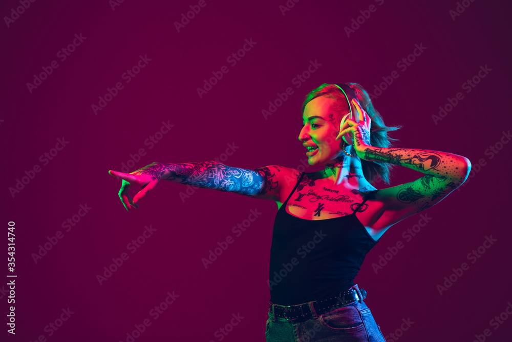 Fototapeta premium Listening to music, dancing with wireless headphones. Portrait of young caucasian woman on purple background in neon light. Human emotions, facial expression, sales, ad concept. Freak's culture.
