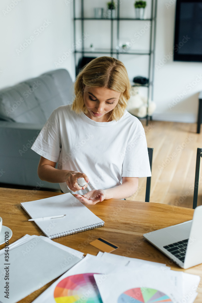 attractive woman holding bottle with hand sanitizer near laptop and credit card on table attractive woman holding bottle with hand sanitizer near laptop and credit card on table