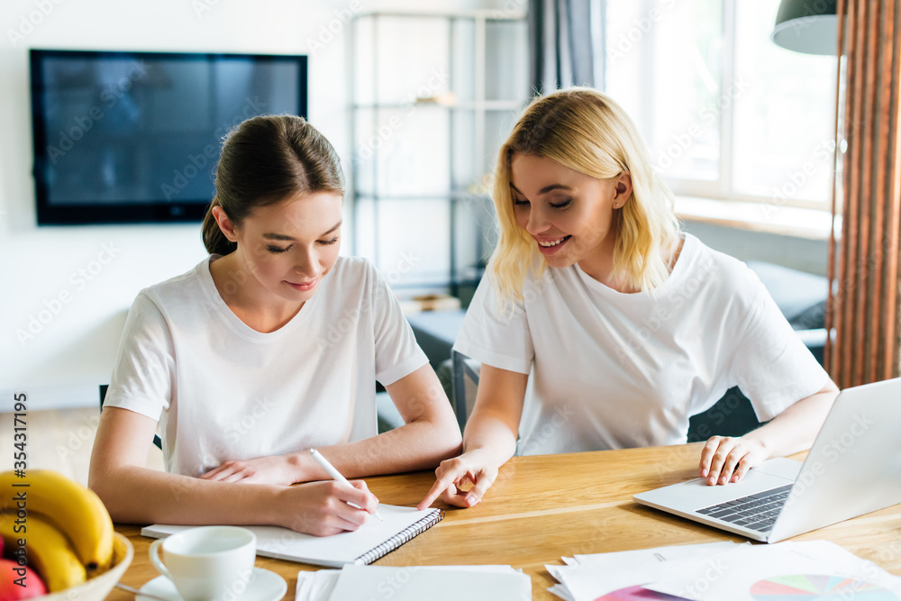 cheerful woman looking at sister writing in notebook neat laptop, online study concept cheerful woman looking at sister writing in notebook neat laptop, online study concept