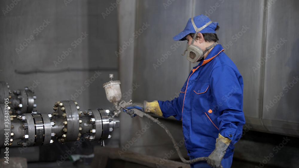 Worker in protective respirator paints oil and gas wellhead tree equipment with spray gun in production plant. Equipment for wellhead conncetion of oil and gas wells. Worker in protective respirator paints oil and gas wellhead tree equipment with spray gun in production plant. Equipment for wellhead conncetion of oil and gas wells.
