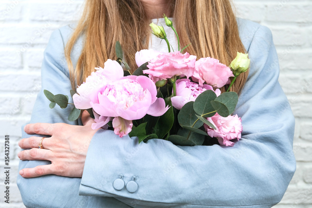 Flower delivery concept of packing flowers. The girl holds pink peonies, against the background of a white brick wall