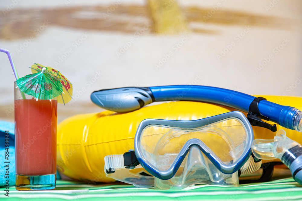 diving mask and snorkel, a glass of fresh tropical juice with cocktail umbrella, a bright yellow rubber ring and a blue towel on a green striped beach mattress. image of sandy beach in the background