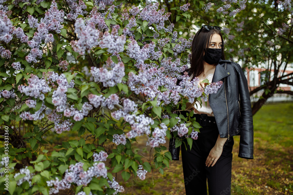Fototapeta premium Girl in a medical mask on a background of blooming lilacs. Black mask. Protection against virus, flu. Coronavirus protection. epidemic of coronavirus. the aroma a tree in the garden Spring allergy