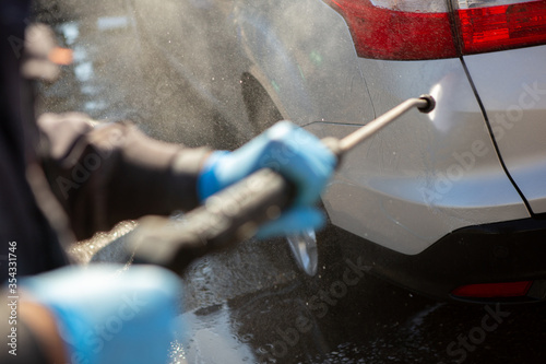A worker in a car wash sprays a gray car with a strong jet of water. The worker wears blue rubber sneakers. Car wash. Copy Space.