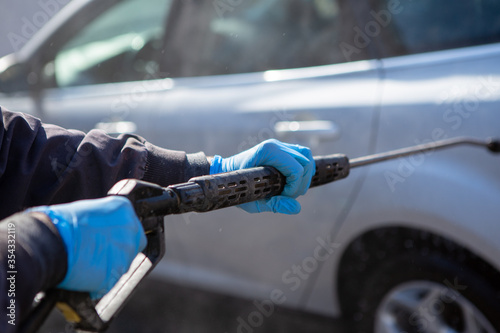 A worker in a car wash sprays a gray car with a strong jet of water. The worker wears blue rubber sneakers. Car wash. Copy Space.