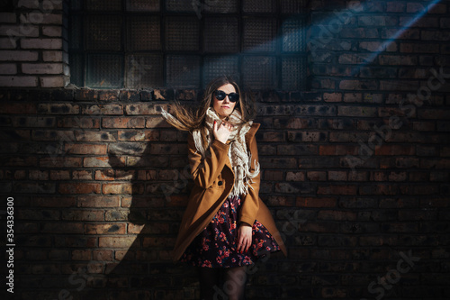 a young beautiful girl in sunglasses and a trench coat stands in a beam of light against a brick wall © Richard