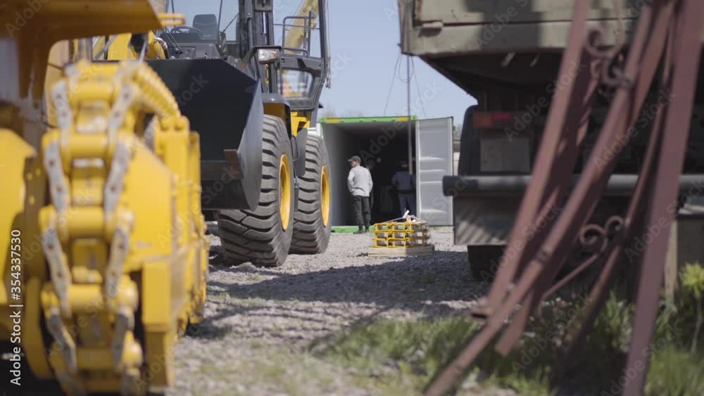 Caucasian construction workers checking equipment in shipping container at the background as heavy industrial machinery standing outdoors at front. Manufature, business, industry.
