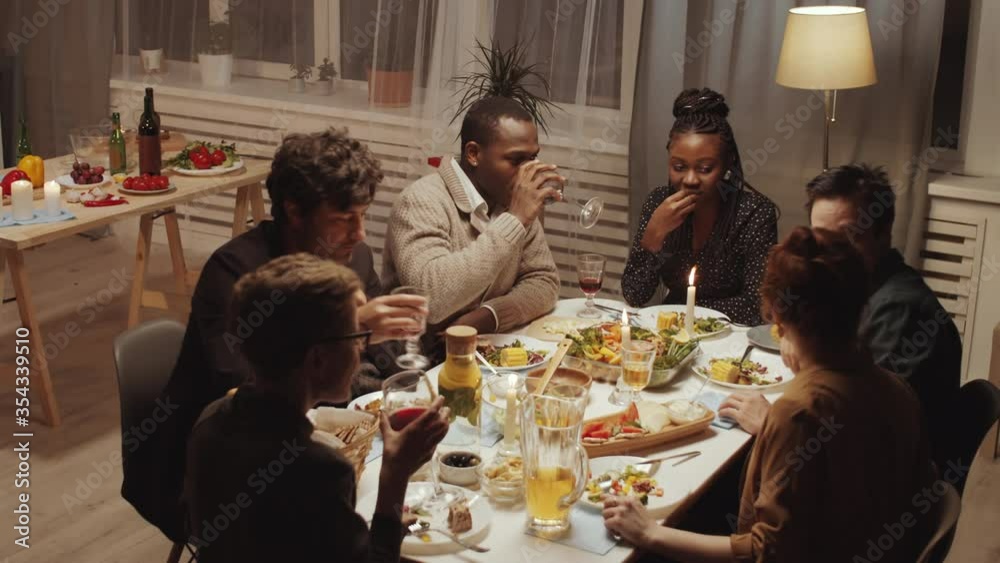 High angle view of group of young multi-ethnic men and women sitting at table together, drinking wine and having discussion at home dinner party High angle view of group of young multi-ethnic men and women sitting at table together, drinking wine and having discussion at home dinner party
