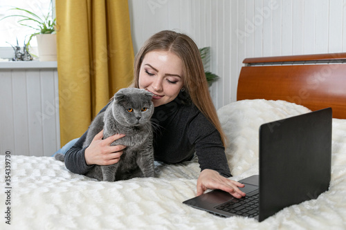 The girl lies on the bed with a laptop. Hugs a gray cat and looks into the laptop. Shopping for pets. Onlan shopping. Online training. Distance learning. We are sitting at home. Purebred cat