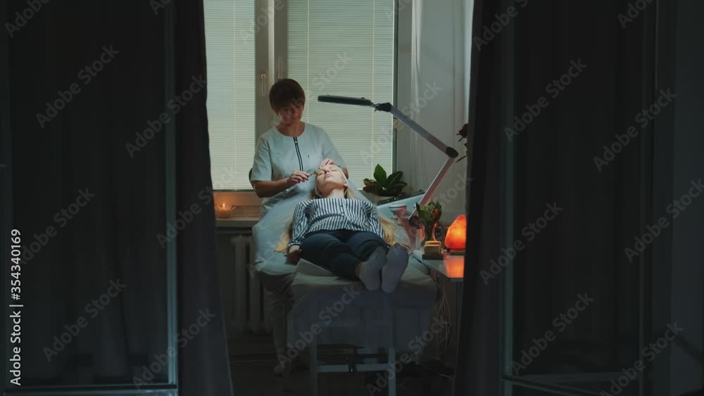 Dolly shot of Beautician making relaxing facial massage with jade roller on woman's face at beauty salon with dim light around and pleasant atmosphere. Dolly shot of Beautician making relaxing facial massage with jade roller on woman's face at beauty salon with dim light around and pleasant atmosphere.
