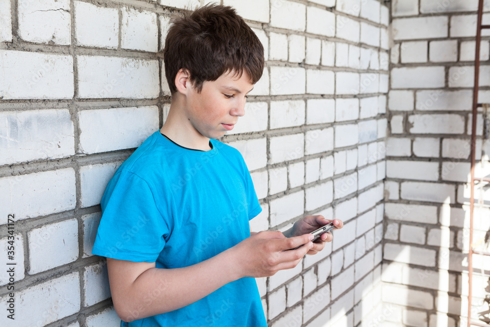 Obraz premium Attractive youngster guy wearing blue t-shirt holding smartphone in hands, browsing social media, leaning brick wall