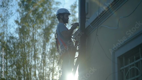 construction worker / laborer secures and tightens a cable standing on ladder on working building site, hard hat and safety equipment, sun flare on camera. Safety first at work concept