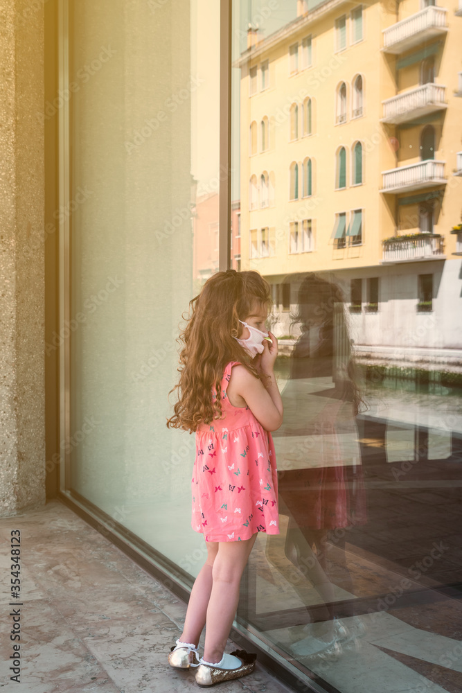 cute little girl wearing a home made surgical mask is looking through a window shop cute little girl wearing a home made surgical mask is looking through a window shop