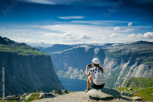 Girl tourist takes pictures. Girl sitting on the edge of a rocky cliff. Extreme tourism. Girl with a backpack. Mountains and fjords of Norway. tourist in a white cap on a cliff looks at the scenery