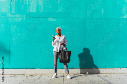 Businesswoman with headphones, standing in front of teal wall, using smartphone