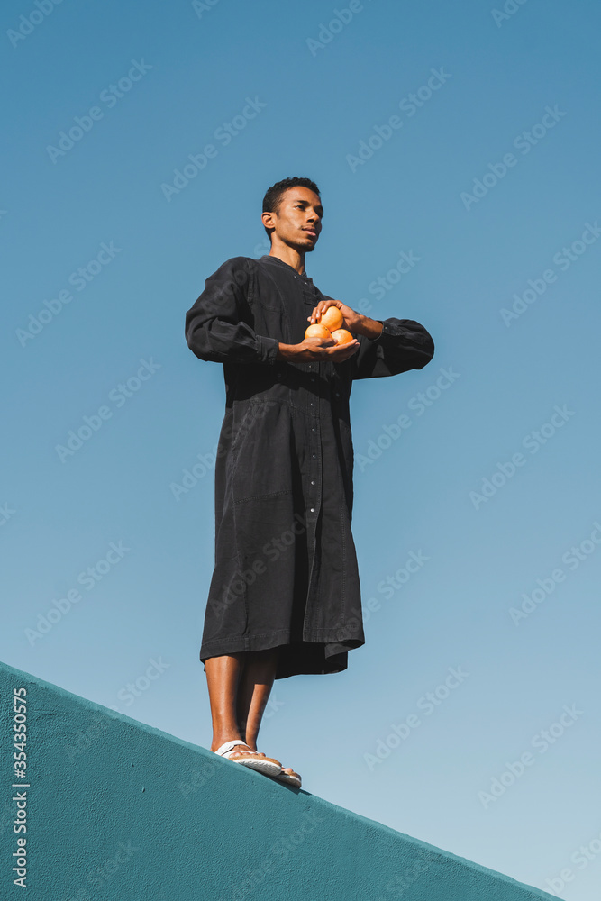 Young man wearing black kaftan standing on blue wall holding oranges Young man wearing black kaftan standing on blue wall holding oranges