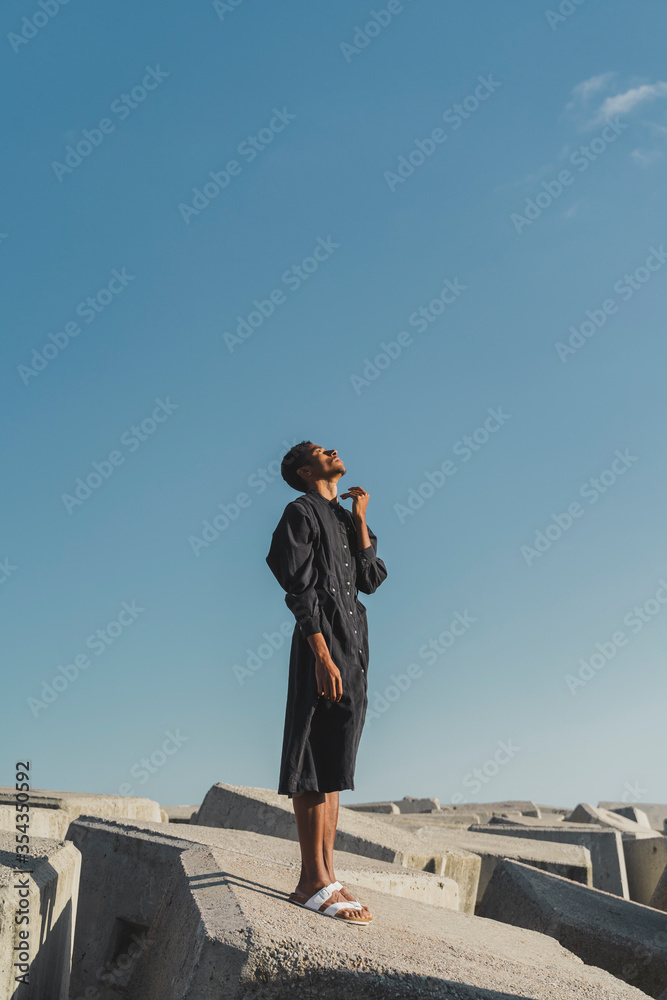 Young man wearing black kaftan standing on concrete blocks under blue sky Young man wearing black kaftan standing on concrete blocks under blue sky