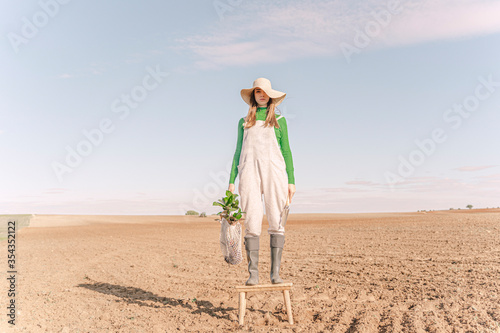 Young woman standing on stool on dry field, carrying plant in a net